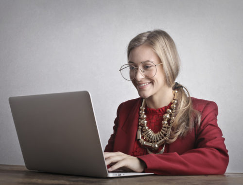Photo of Smiling Woman in a Red Coat and Glasses Using a Laptop
